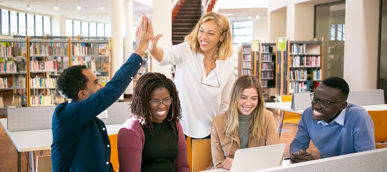 A group of four young teachers and a school leader gather around a laptop in a library, smiling, as the leader gives one of the teachers a high five.