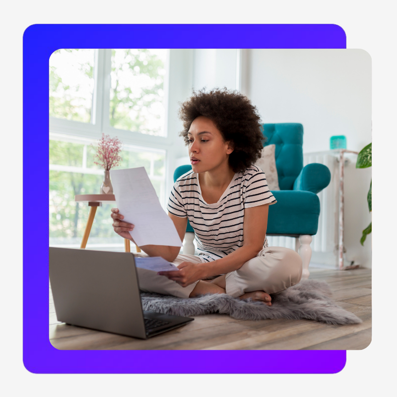 A Black woman with short curly hair, a striped shirt, and khaki pants sits crosslegged on the floor looking at paper work. She has a laptop open in front of her, behind her is her living room, with a comfy chair, and windows letting filtered light in