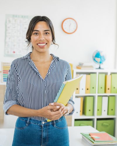 This photograph captures a woman standing in a classroom, holding a yellow folder and wearing a blue-and-white striped blouse. Behind her, shelves filled with green binders and a globe suggest a well-organized educational setting.